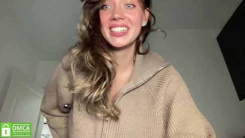 Media: A video of a smiling, light-skinned woman with wavy, shoulder-length brown hair, wearing a beige ribbed hoodie. She has a soft, rounded face, small ears, and is indoors with a white door and black wall in the background.