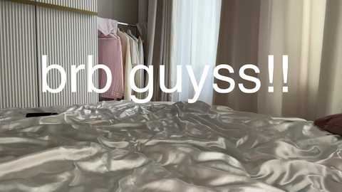 Media: Video of a messy, unmade bed with wrinkled white sheets in a dimly lit bedroom. Clothes hang on a closet door and sheer curtains partially cover a window, with text overlaying \"brrb guys!!\