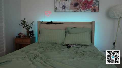 Media: Video of a bedroom with a green bedspread, two pillows, wooden headboard, framed floral painting, a nightstand, and a white floor lamp.