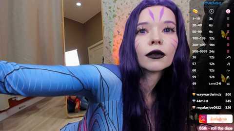 Media: Video of a young woman with long, purple hair and purple face paint, wearing a blue bodysuit. Background shows a bedroom with floral wallpaper, a door, and a mirror. Streaming overlay on right.