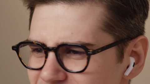 Media: A close-up video of a young man with fair skin, short brown hair, wearing black rectangular glasses, white earbuds, and a subtle smile. The background is a plain, neutral beige.