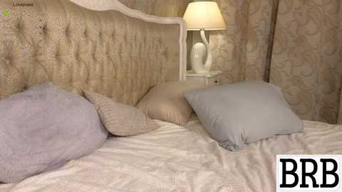 Media: Video of a plush, tufted beige headboard bed with a cream quilt, two pillows, a nightstand with a lamp, and a floral wallpaper background.