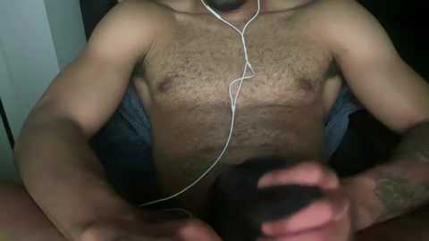 Media: A video of a shirtless, light-skinned man with a hairy chest, wearing headphones, sitting on a black surface, holding a black object.