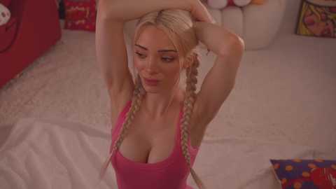 Media: Video of a blonde woman with large breasts, wearing a pink tank top, stretching her arms above her head in a playful, indoor setting with colorful pillows.