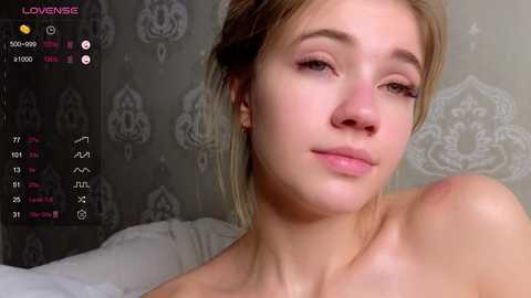 Media: Video of a young Caucasian woman with fair skin and blonde hair, smiling slightly, with a soft-focus background featuring patterned wallpaper.