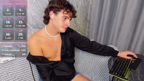 Media: Video of a young man with curly dark hair, wearing a black off-shoulder shirt, white pearls, and wireless earbuds, seated at a desk with a gaming keyboard.