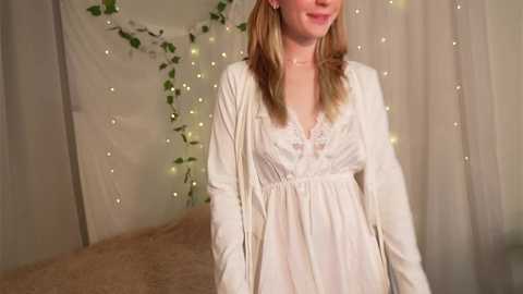 Media: Video of a smiling, fair-skinned woman with long blonde hair, wearing a white, lace-trimmed nightgown, standing in a cozy bedroom with beige curtains and fairy lights.