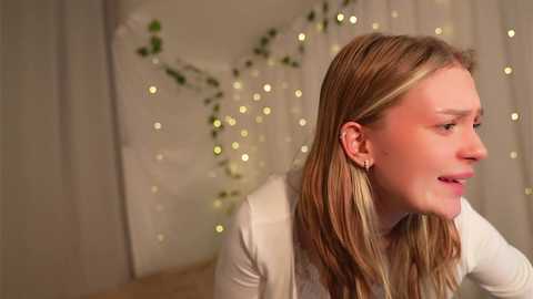 Media: Video of a young, fair-skinned woman with long blonde hair, wearing a white top, looking to the right. Background features soft, bokeh lights and green ivy.