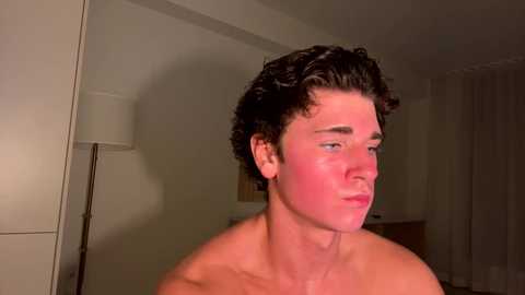 Media: Video of a shirtless, light-skinned, curly-haired young man with flushed cheeks, possibly from embarrassment, standing in a dimly lit, sparsely furnished room with a lamp and a bed in the background.