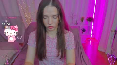 Media: Video of a young woman with long brown hair, wearing a pink floral top, seated in a room with pink lighting, background features a Hello Kitty decoration and a potted plant.