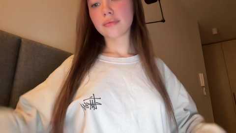 Media: Video of a young woman with long, straight, auburn hair, fair skin, and a neutral expression, wearing a loose, white hoodie with a black logo, standing indoors against a beige wall.