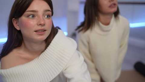 Media: Video of two young women with fair skin and straight brown hair, wearing white knit sweaters, looking thoughtful. Background features a modern, dimly-lit room with blue lighting.