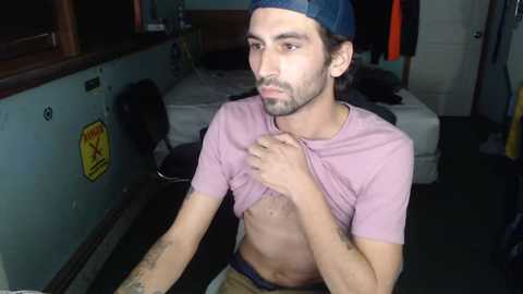 Media: Video of a young man with dark hair and beard, wearing a blue cap and pink shirt, lifting shirt to reveal bare chest in dimly lit room with bed, posters, and warning signs.