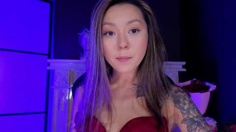 Media: Video of a young, light-skinned woman with long, straight brown hair, wearing a strapless red dress, standing in a dimly lit room with purple lighting. She has a tattoo sleeve on her right arm.