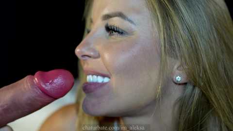 Media: Video of a blonde woman with fair skin, wearing small earrings, smiling while licking a circumcised, erect penis, against a dark background.