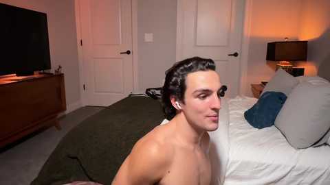 Media: Video of a shirtless, fair-skinned man with dark hair, lying on a bed in a dimly lit, cozy bedroom with a flat-screen TV, wooden dresser, and a lamp on the nightstand.