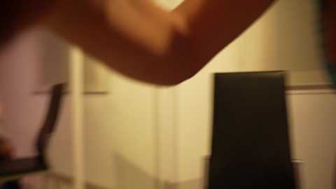 Media: A blurred video of a person's arm, hand, and shoulder, with a black box and white walls in the background. The image is out of focus, with a warm, ambient lighting.