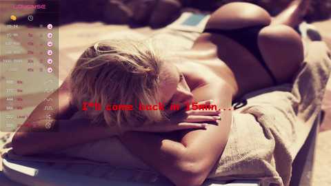 Media: Video of a blonde woman lying face down on a beach towel, wearing black thong bikini, text overlay, sunlit background.