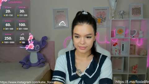 Media: Video of a young woman with fair skin, dark hair in a ponytail, wearing a navy blue and white sailor-style top, sitting in a pink room with a stuffed elephant, pink lights, and various decorations.