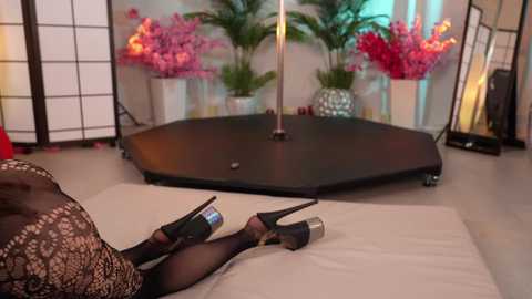 Media: Video of a woman lying on a beige couch, legs crossed, wearing black fishnet stockings and high heels, with a pole dance pole in the background, surrounded by vibrant pink flowers and green plants.