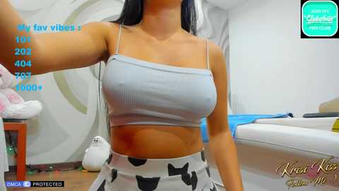 Media: A video of a woman with medium skin tone, wearing a light blue ribbed crop top and black cow-patterned shorts, in a modern, minimalist bedroom.