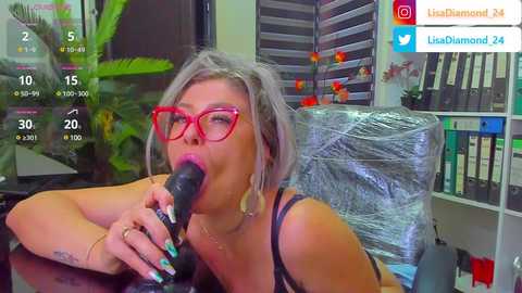 Media: A video of a woman with platinum blonde hair, wearing red glasses, black bra, and blue nail polish, sucking a black dildo while seated at a desk. Background includes office items and social media icons.