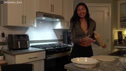 Media: A smiling Asian woman in a black zip-up jacket and black pants bakes in a modern kitchen with white cabinets, black appliances, and a tiled backsplash.