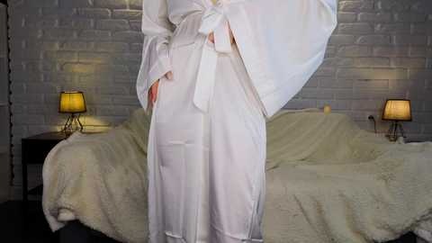 Media: A video of a person in white bathrobe, standing in front of a beige bed with a white blanket, white brick wall background, two bedside lamps, and a lampshade.