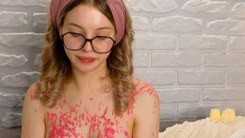 Media: Video of a young woman with light skin, wavy blonde hair, and large black glasses, wearing a pink headband, covered in red paint. She stands in front of a white brick wall, holding a yellow candle.