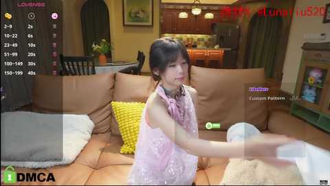 Media: A video of a young Asian woman in a pink dress, playing a video game on a brown leather couch in a modern living room with wooden cabinets and a kitchen in the background.