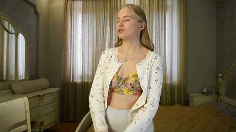 Media: A video of a blonde woman with fair skin, wearing a white floral-patterned bra and matching pants, standing in a dimly lit bedroom with beige curtains and a cream-colored dresser.