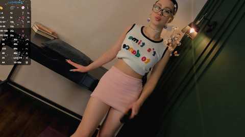 Media: A video of a young woman with glasses and a white crop top featuring colorful letters, wearing a pink mini skirt, standing in a dimly lit room with wooden floors and a green wall.