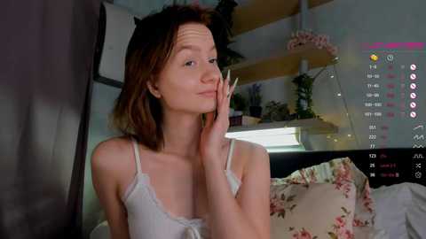 Media: Video of a young, slender, fair-skinned woman with shoulder-length brown hair, wearing a white lace camisole, sitting on a bed with floral pillows. The background shows a cluttered room with plants and shelves.