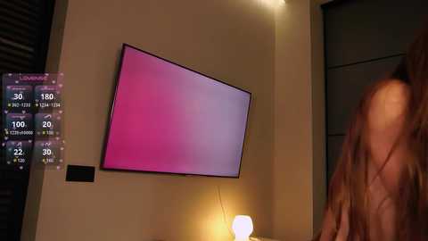 Media: Video of a modern room with a large, flat-screen TV displaying a pinkish hue. The wall is beige, and a digital clock displays time. A person with long hair stands in the right background, partially out of focus.