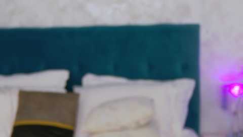 Media: A blurry video of a bed with white pillows and a teal headboard, partially visible. The background features a purple light source on the right and a beige wall.