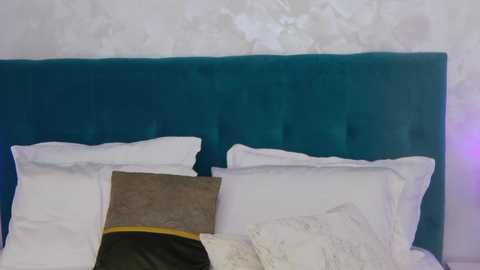 Media: Video of a neatly made bed with a teal velvet headboard, featuring white pillows, a brown pillow with a yellow trim, and white sheets, set against a textured white wall.