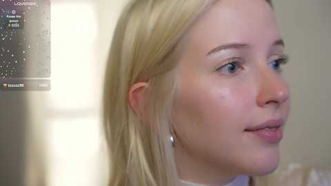 Media: Video of a fair-skinned, blonde woman with straight hair, wearing a white top, looking to the right with a neutral expression. Background is out of focus.