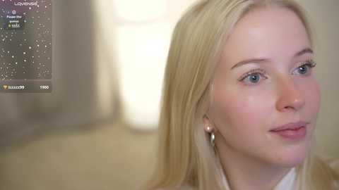 Media: Video of a young, fair-skinned blonde woman with straight hair, wearing a white blouse, looking thoughtfully to the right. Background is blurred, suggesting an indoor setting.