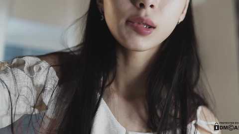 Media: Video of an Asian woman with long black hair, wearing a white off-shoulder top, with lips slightly parted, looking pensive. Background is blurred, featuring a beige wall and a blue object.