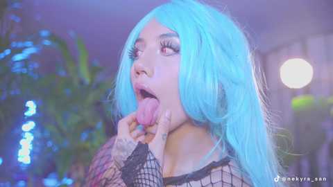 Media: Video of a young woman with long, pastel blue hair and a fishnet top, licking her fingers with a playful expression, in a dimly lit room with blurred greenery in the background.