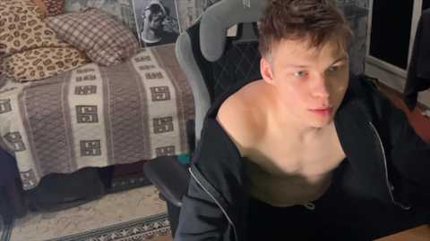 Media: Video of a young Caucasian man with short brown hair, wearing a black shirt, sitting on a gaming chair in a cluttered room with a bed and patterned carpet.