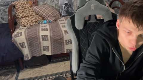 Media: Video of a young man in a black hoodie sitting on a gaming chair, looking at a phone. A messy bed with leopard-print bedding and a dog visible in the background.