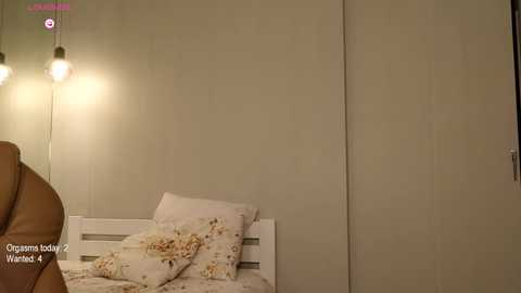 Media: Video of a minimalist bedroom with beige walls, a white wooden headboard, a brown leather chair, and two decorative pillows with floral patterns. A single hanging light fixture casts a warm glow.