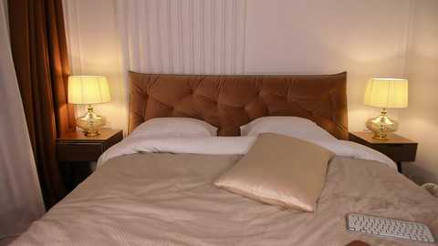 Media: Video of a modern, cozy bedroom with a tufted brown headboard, beige bedspread, two white pillows, two nightstands with gold lamps, beige curtains, and a white laptop on the bed.