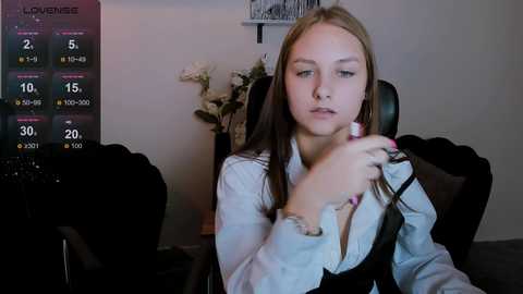 Media: Video of a young woman with long brown hair, wearing a white blouse and black vest, holding a comb, in a dimly lit room with a \"London\" weather forecast screen and flowers.