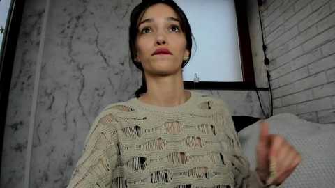 Media: Video of a woman with fair skin, dark hair, and light-colored knit sweater in a dimly lit room with gray marbled walls and a brick wall.