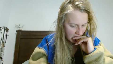 Media: A video of a blonde woman with fair skin, wearing a blue and yellow sweater, sitting in a bedroom with a wooden dresser and a decorative deer head. She's yawning, her hand near her mouth.