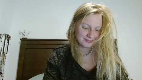 Media: Video of a smiling, blonde, fair-skinned woman with long hair in a dark, patterned top, standing in a minimalist bedroom with a wooden headboard.