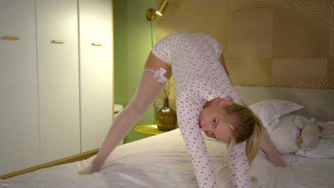 Media: Video of a slender, blonde Caucasian woman in a white polka-dotted leotard and thigh-high stockings, performing a split on a bed in a modern, green-walled bedroom with a patterned headboard.