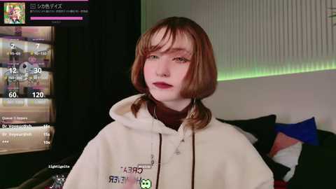 Media: Video of a young woman with short, auburn hair, wearing a cream hoodie, sitting in a dimly lit room with gaming setup, including a monitor and green backlit panel.